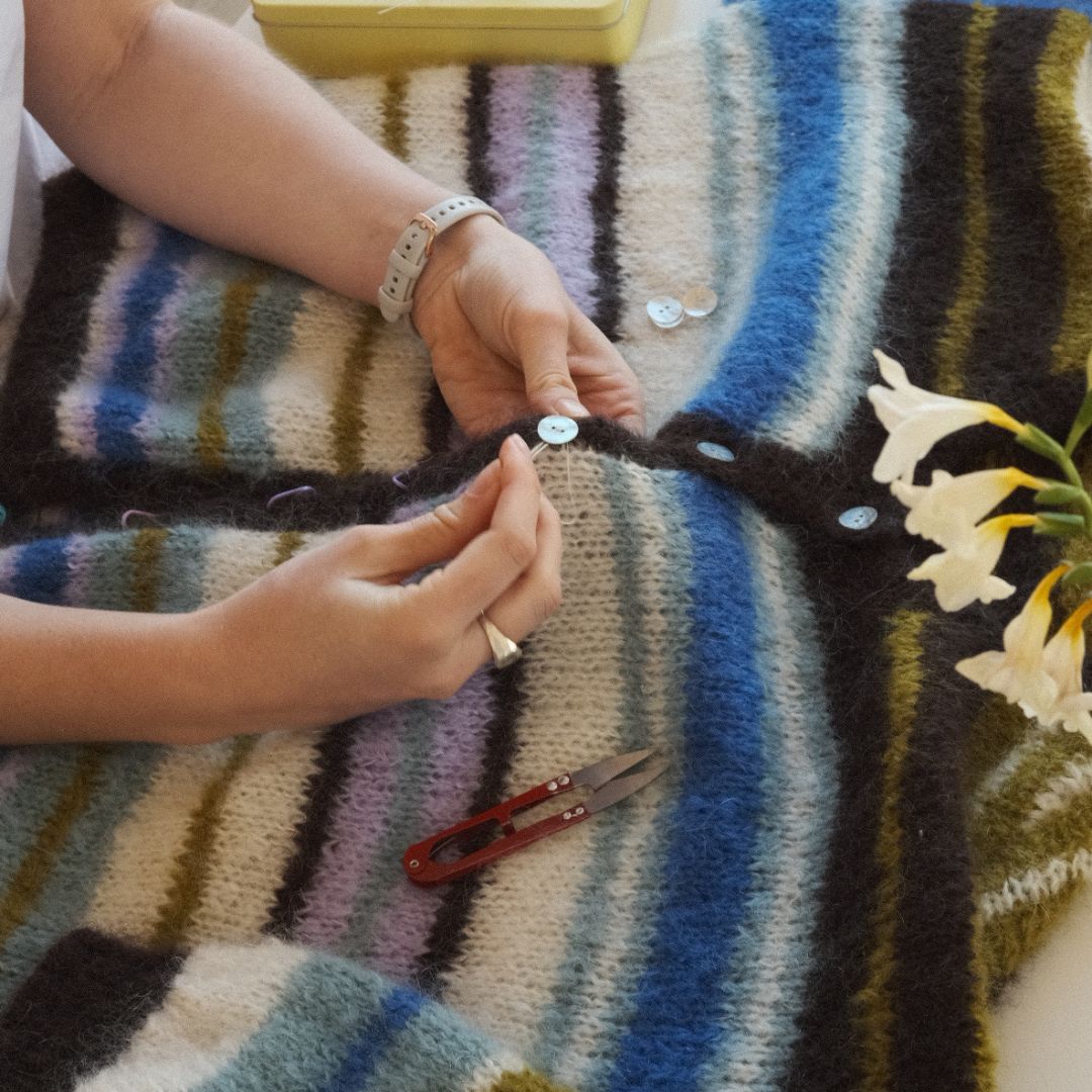 Person sewing buttons onto a striped fabric with a pair of scissors and flowers nearby.