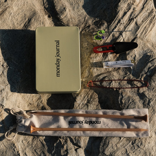 Collection of items including a 'mondayjournal' tin, a knife, and wooden tools on a rocky surface.