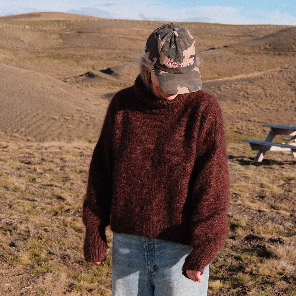 Person wearing a brown jacket and light blue jeans standing on a rocky surface with a clear sky.